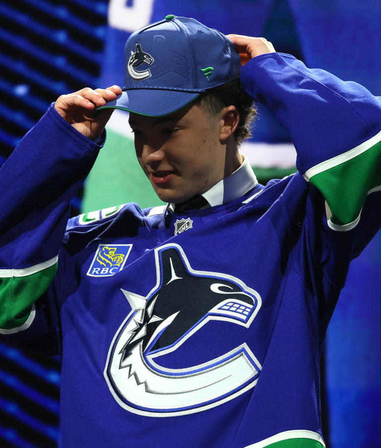 Caleb Malhotra wearing a blue hockey jersey and cap with a logo, standing against a blurred background.